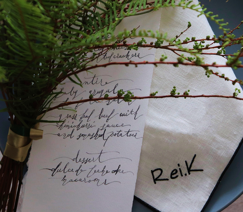 手書きのメニューカードと『Rei.K』と刺繍された白いナプキン、添えられたグリーンの小枝